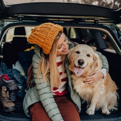 Photo of a young smiling woman and her dog sitting the trunk of a car, taking a short break during their road trip.